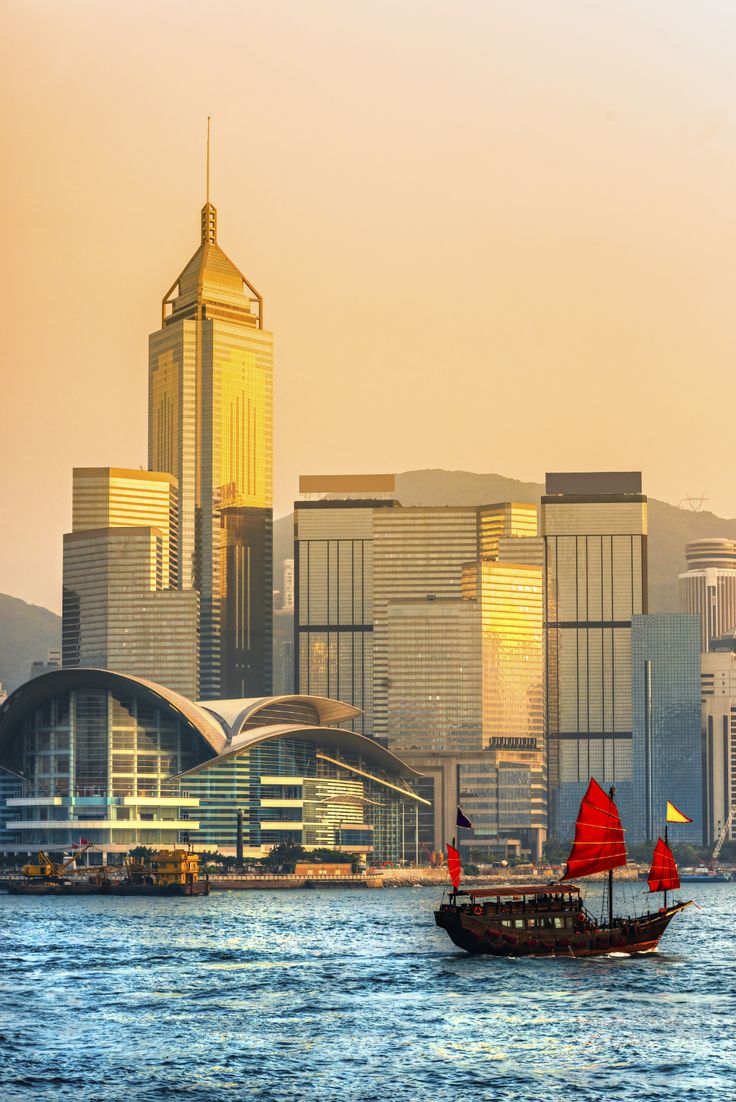 a boat with red sails in the water next to tall buildings and a city skyline