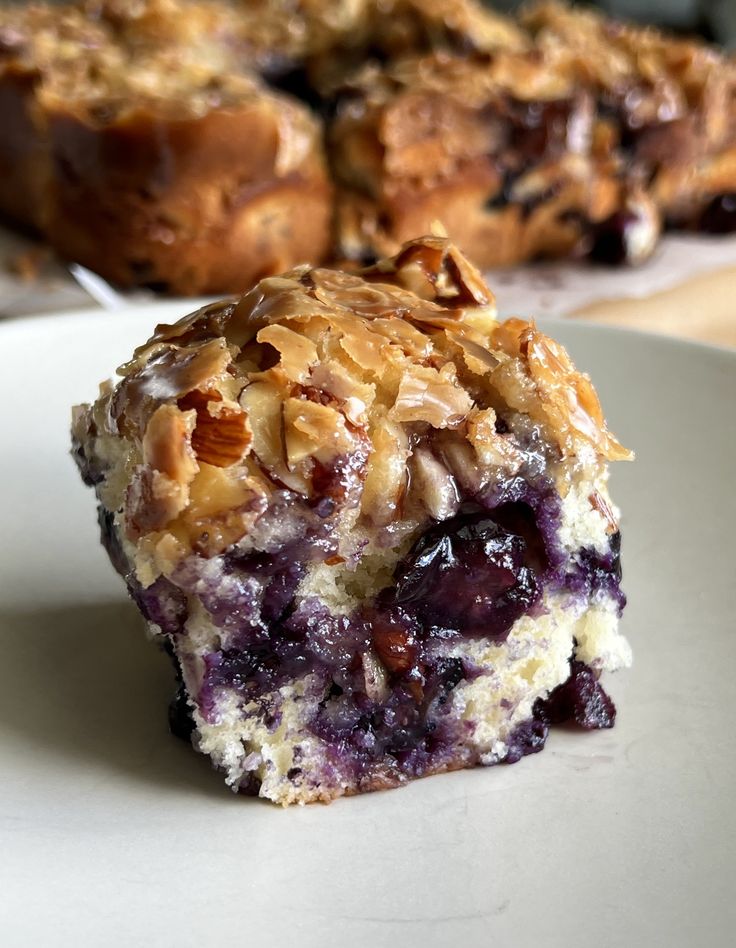 a piece of blueberry muffin on a plate with another muffin in the background