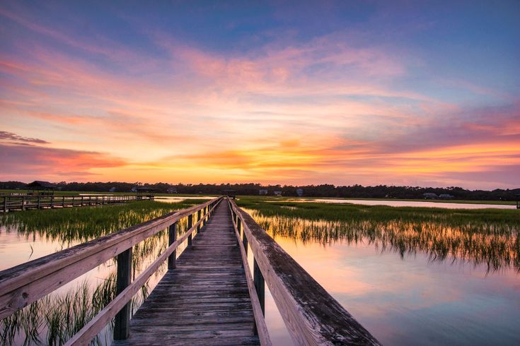 a wooden bridge over water at sunset with grass in the foreground and clouds in the background