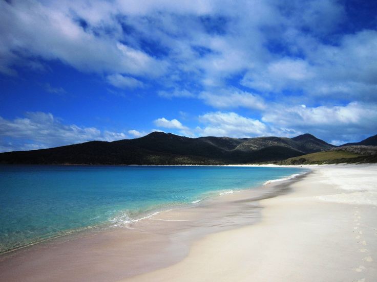 a sandy beach with blue water and mountains in the background