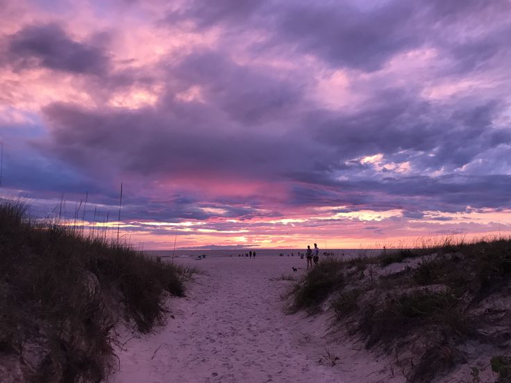 people are walking on the beach as the sun goes down in the distance with purple and pink clouds