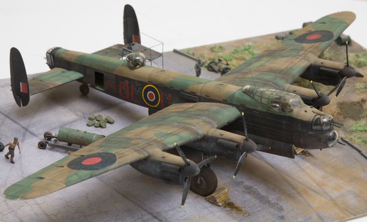 two model airplanes are on a table with people standing around and one plane is camouflaged