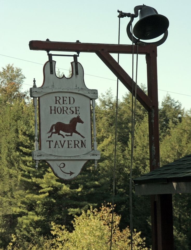 a red horse tavern sign hanging from the side of a wooden building with trees in the background