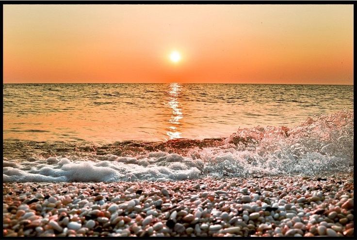 the sun is setting over the ocean with small rocks in the foreground and waves crashing on the shore