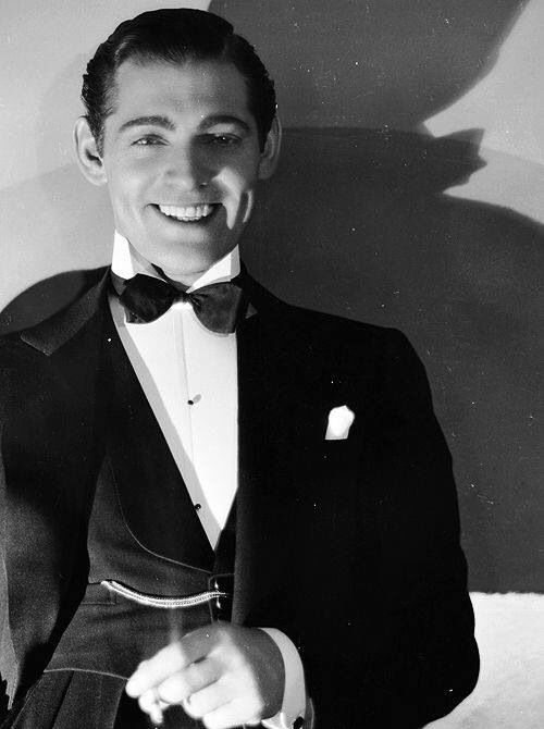 a man in a tuxedo and bow tie smiles at the camera while holding his hand out