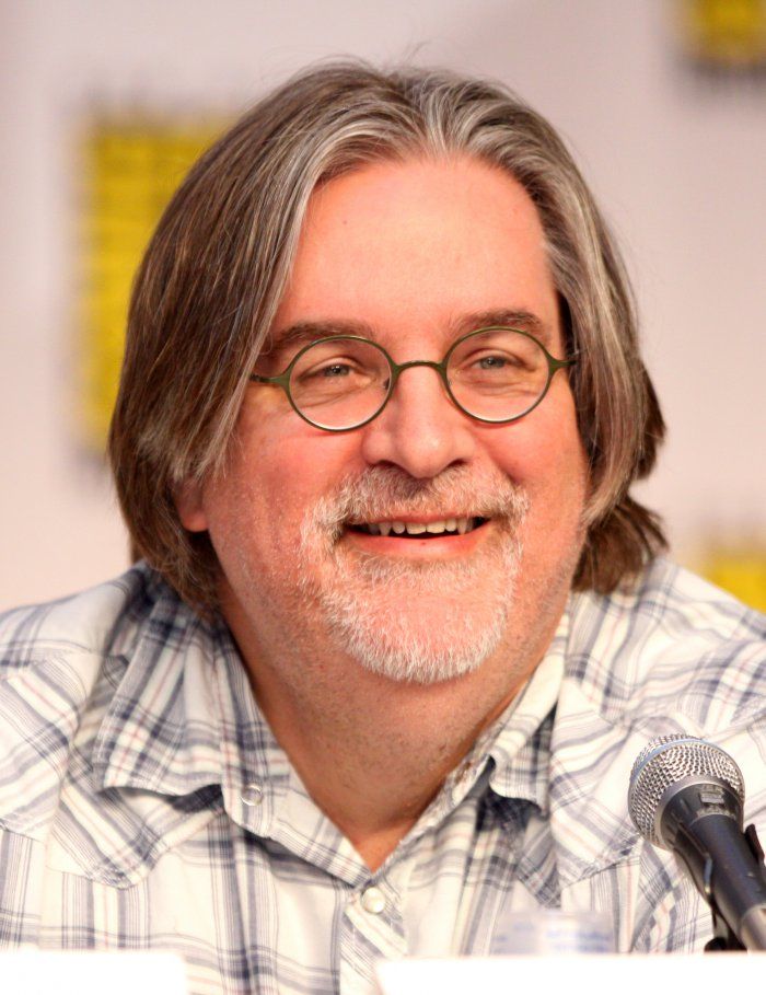 an older man with glasses and a plaid shirt smiles while sitting in front of a microphone