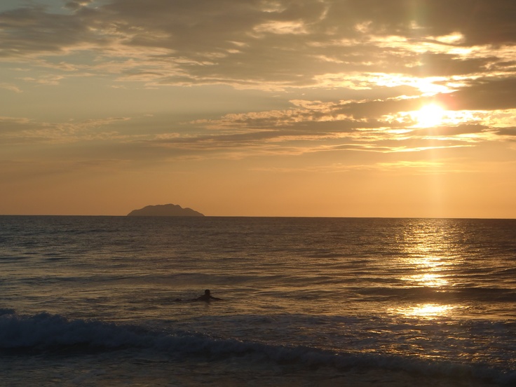 the sun is setting over the ocean with surfers in the water and an island in the distance