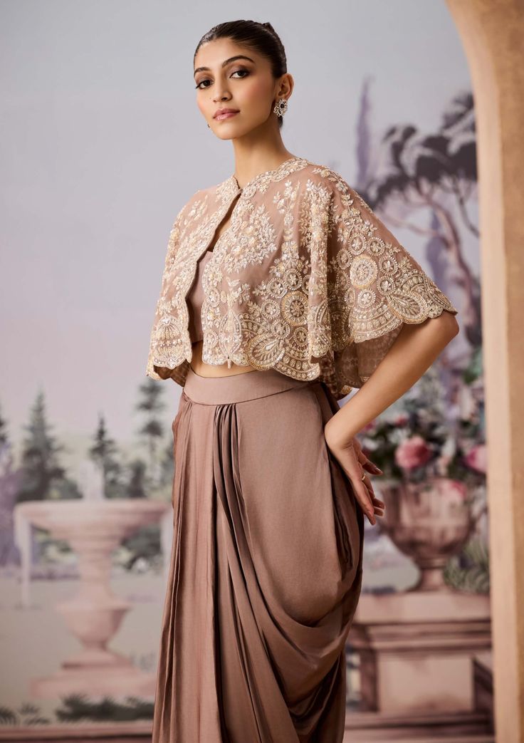 Featuring a luxurious golden satin dhoti skirt paired with a stunning fully hand-embroidered cape. The cape is adorned with exquisite cutdana, sequins, and beadwork, adding an opulent touch to the ensemble. Complemented by a chic bralette blouse, this outfit effortlessly marries modern aesthetics with traditional craftsmanship, making it a captivating choice for special occasions or celebrations. Semi-stitched Draped Lehenga For Eid, Festive Draped Traditional Wear With Resham Embroidery, Eid Draped Semi-stitched Lehenga, Designer Draped Lehenga With Resham Embroidery, Traditional Unstitched Draped Sets, Traditional Wear With Zari Work And Cape Sleeves, Embroidered Sharara With Cape Sleeves For Festive Occasions, Festive Embroidered Palazzo Set With Cape Sleeves, Navratri Traditional Wear With Dupatta And Cape Sleeves