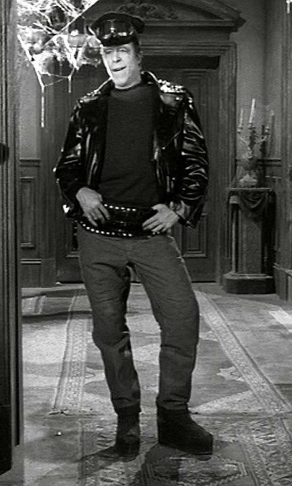 the man is wearing a leather jacket and standing in front of a door with his hands on his hips