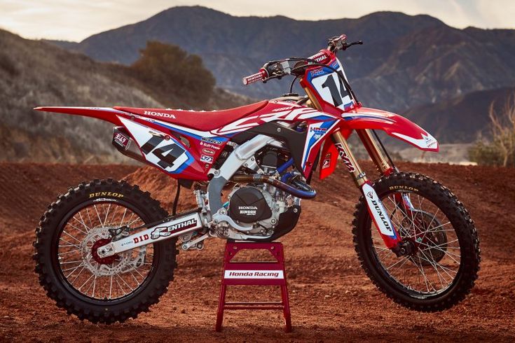 a red and white dirt bike parked on top of a dirt field with mountains in the background