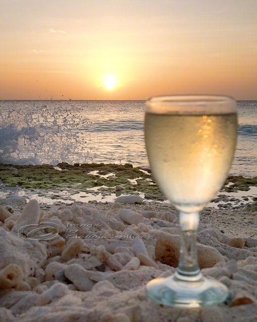 a glass of wine sitting on top of a sandy beach next to the ocean at sunset