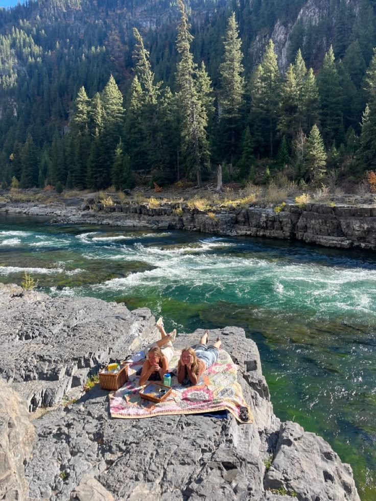 two people are laying on the rocks by the river while having a picnic with their dog