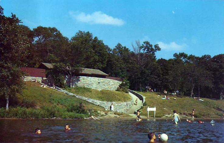 many people are swimming in the water near a stone wall and grassy hill with trees