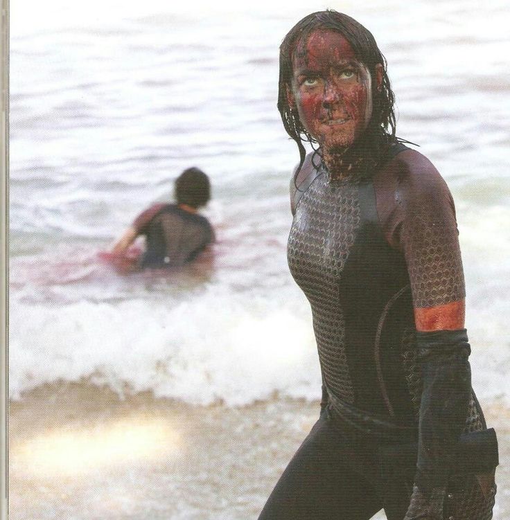 a woman in wetsuit standing on beach next to surfboard with other people in water behind her