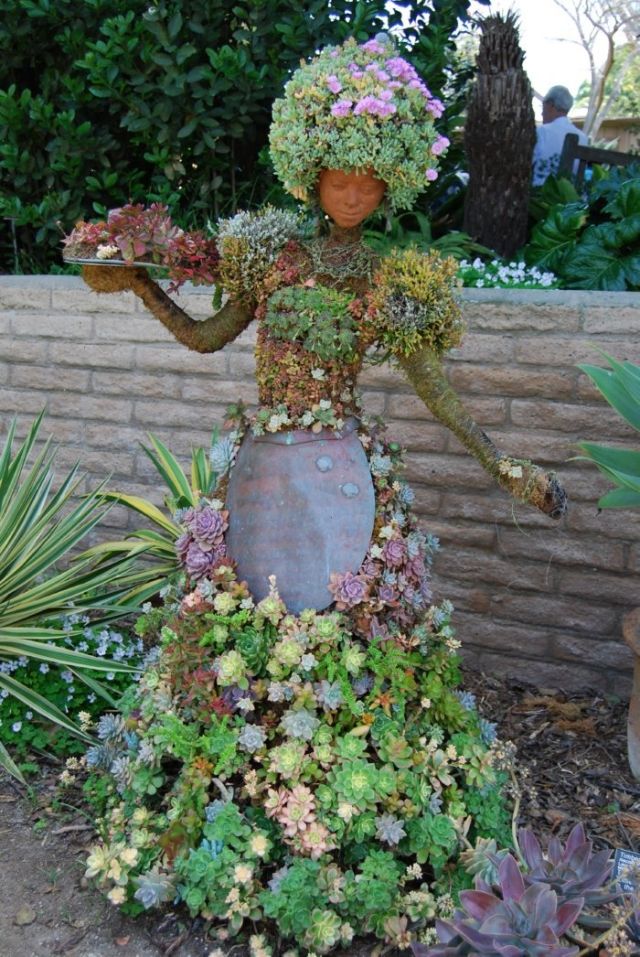 a statue made out of plants and flowers in front of a brick wall with a woman holding