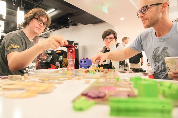 two men playing board games at a table with other people in the background looking on