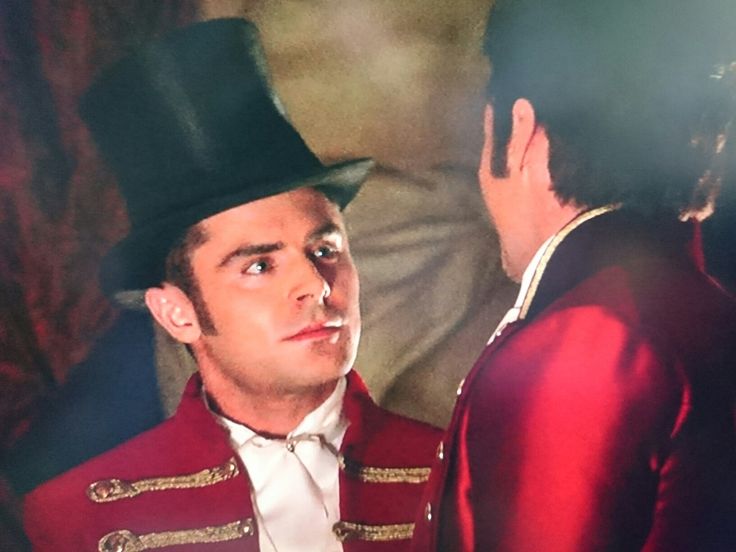 a man in a red jacket and top hat talking to another man wearing a white shirt