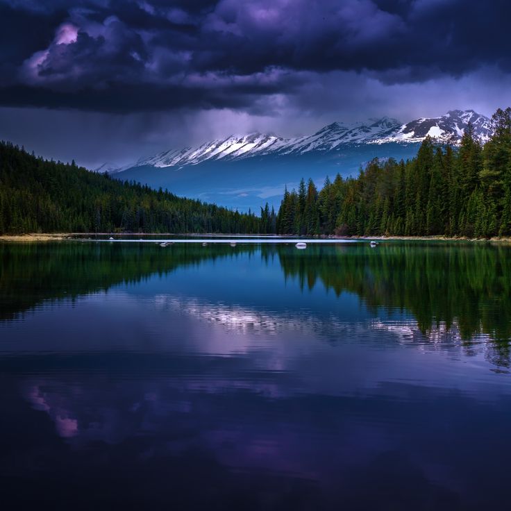 a lake surrounded by trees and mountains under a cloudy sky with snow capped mountain in the distance