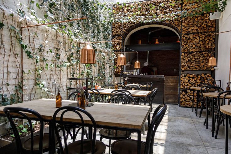 an outdoor dining area with tables, chairs and wood stacked up on the wall behind it