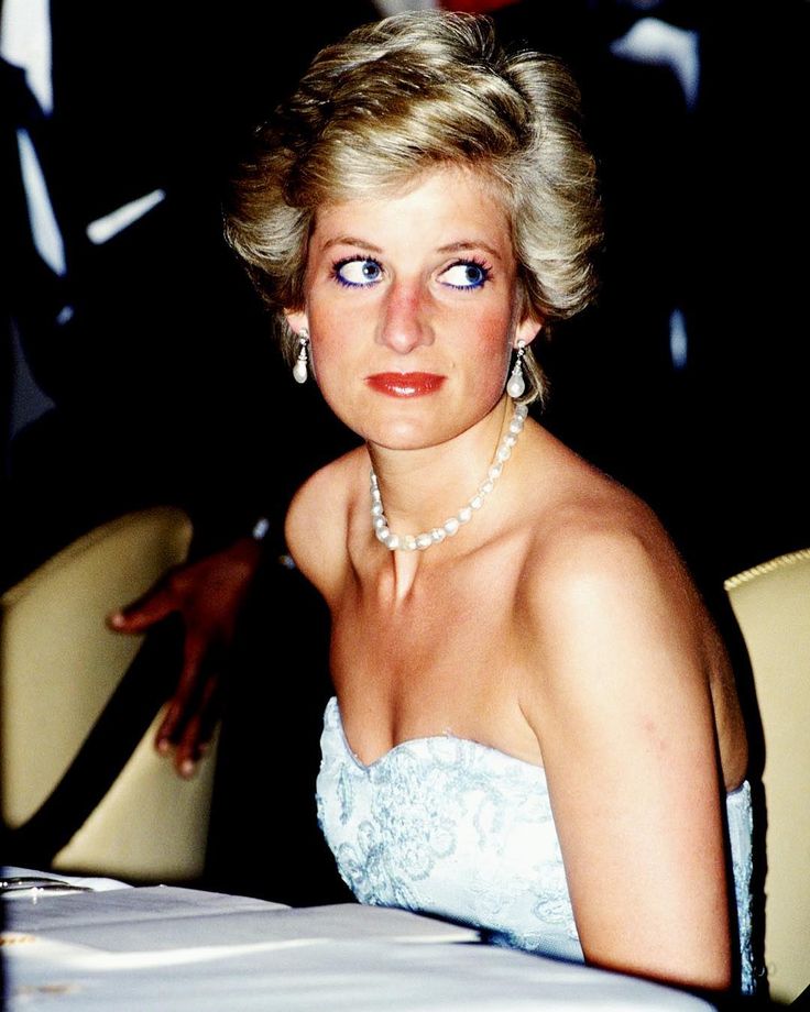 a woman sitting at a table wearing a white dress and pearl necklace with her hand on her shoulder