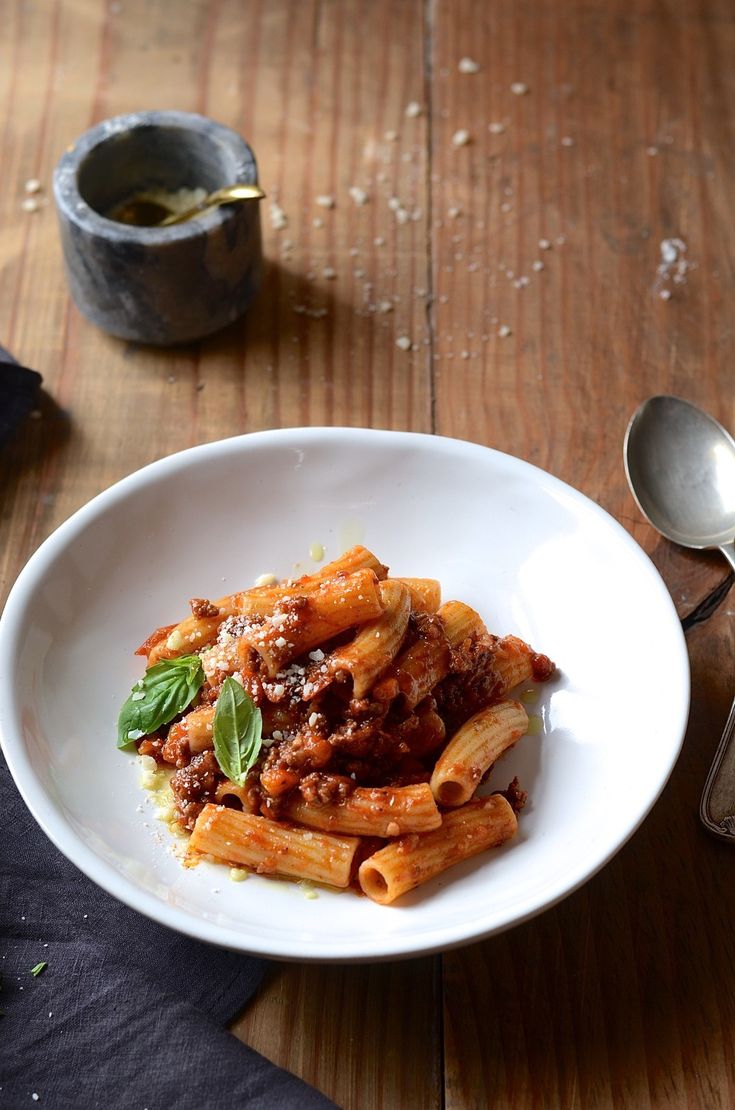 a white bowl filled with pasta and sauce on top of a wooden table next to a spoon