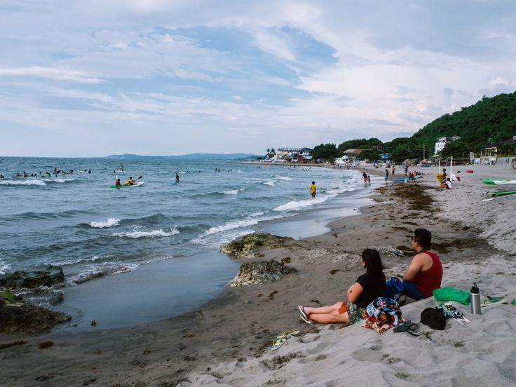 two people are sitting on the beach looking out at the ocean while others swim in the water