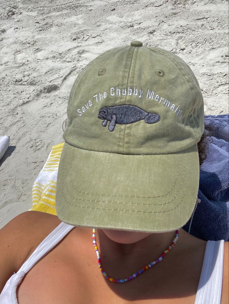 a woman sitting on the beach wearing a green hat with a fish embroidered on it