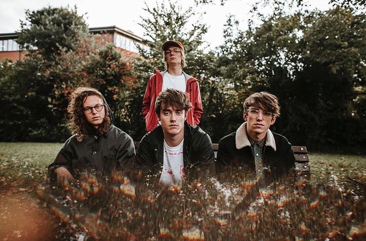 four young men are sitting on a bench in the middle of some grass and trees