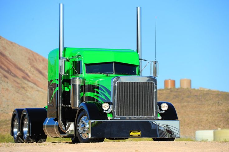 a green semi truck parked on the side of a dirt road with mountains in the background