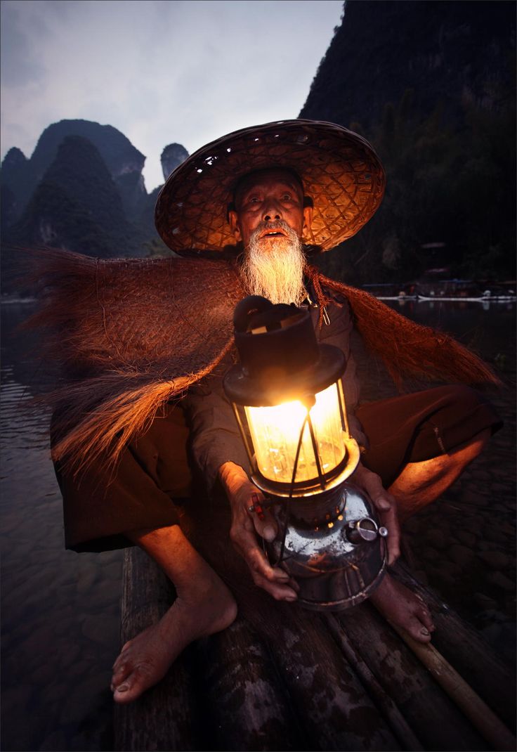 a man sitting on top of a wooden dock holding a lantern in his hand and wearing a hat