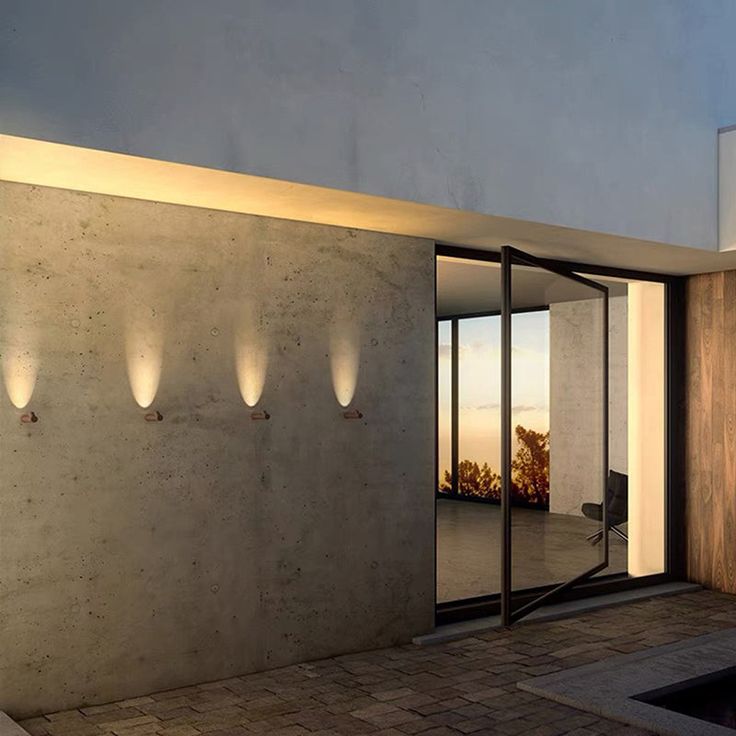 the outside of a modern house with lights on it's walls and doors open