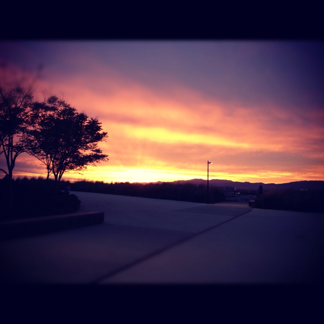 the sun is setting behind some trees and hills in the distance, as seen from an empty parking lot