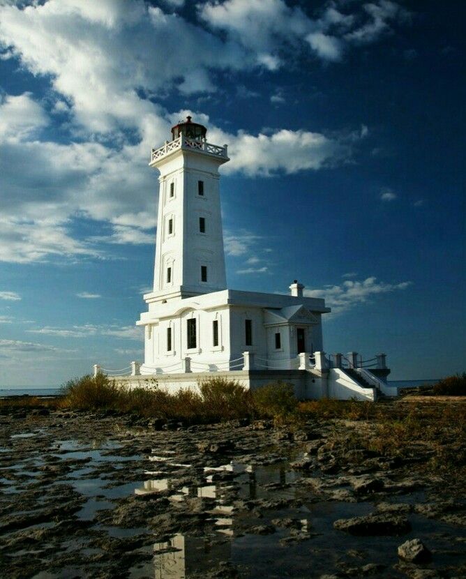 a white lighthouse sitting on top of a rocky beach next to the ocean under a cloudy blue sky