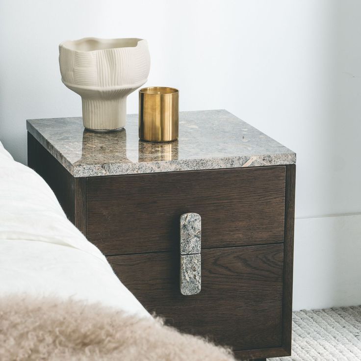 a white vase sitting on top of a table next to a wooden drawer and bed