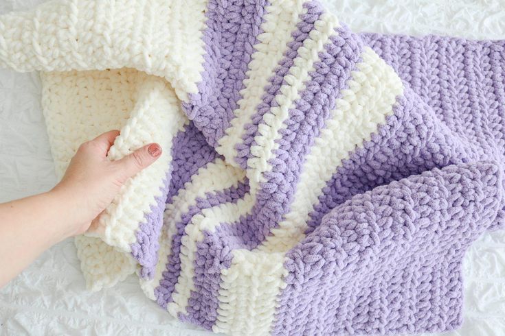 someone is crocheting a blanket on top of a white bed sheet with purple and white stripes