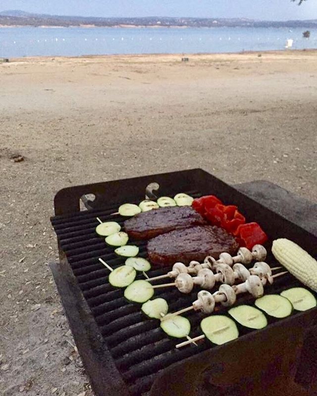 a bbq grill with vegetables and meat cooking on it near the water's edge