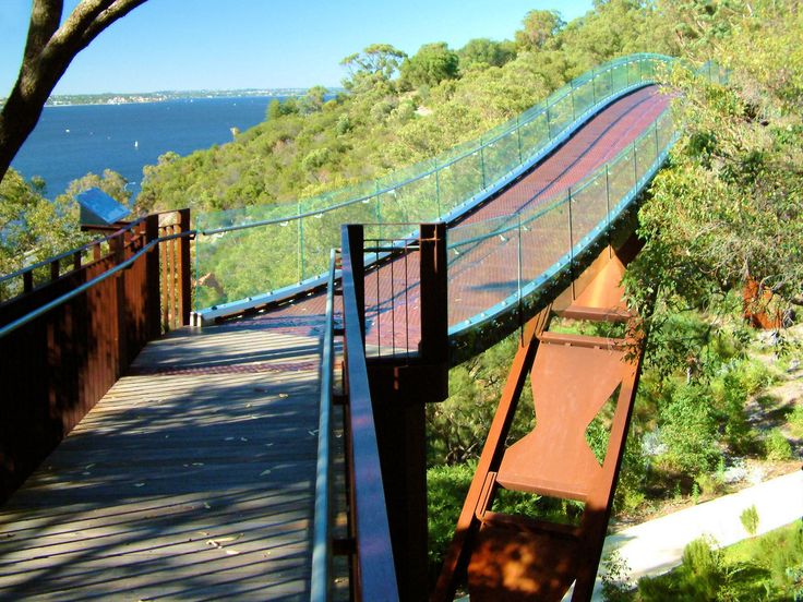 a wooden walkway that is next to some trees and water in the distance, with a bridge going over it