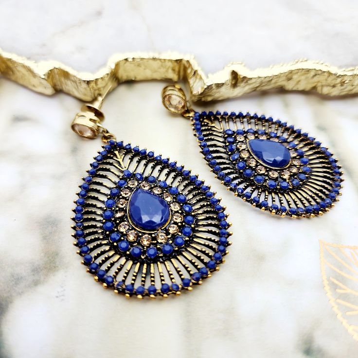 Captivating in its royal essence, this jewelry set features a striking combination of deep blue stones and gold-toned metalwork. The necklace, with its simple yet elegant golden bead chain, leads to a pendant boasting a rich blue stone at its core, encircled by a halo of darker blue crystals. The showstoppers are the chandelier earrings, presenting an intricate peacock feather-inspired design with a teardrop blue stone centerpiece, framed by layers of fine, dark blue crystals that create a drama Antique Gold Bohemian Jewelry For Party, Bohemian Antique Gold Jewelry For Party, Blue Gold Plated Jewelry For Party, Sapphire Metal Jewelry For Party, Elegant Jeweled Sapphire Jewelry, Elegant Blue Stone Jewelry, Elegant Antique Gold Teardrop Jewelry, Elegant Sapphire Jeweled Jewelry, Sapphire Party Jewelry