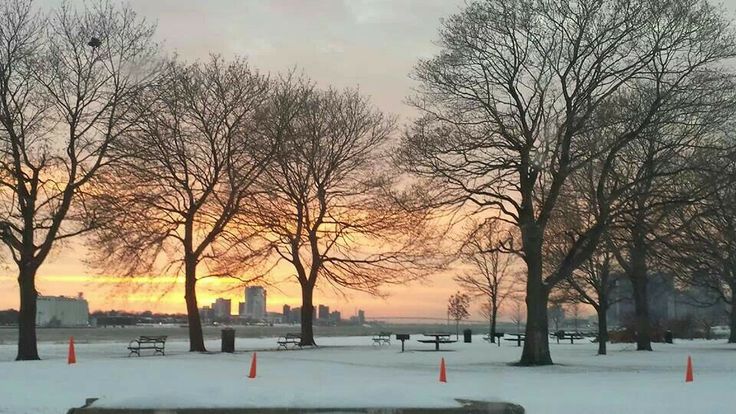 the sun is setting behind some trees and benches in the snow covered park with orange cones