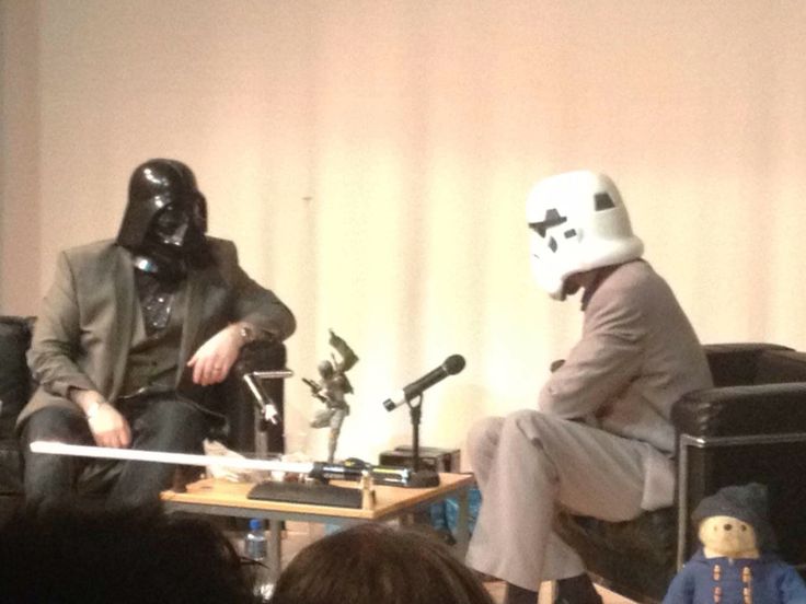 two people sitting on chairs in front of microphones with one person wearing a darth vader mask