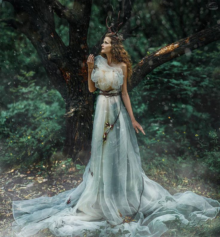 a woman in a white dress standing next to a tree with deer antlers on her head