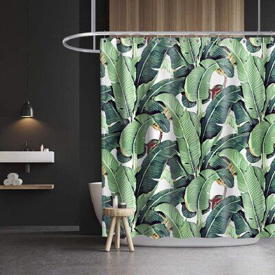 a green shower curtain with banana leaves on it in a dark bathroom area, next to a white toilet and wooden stool