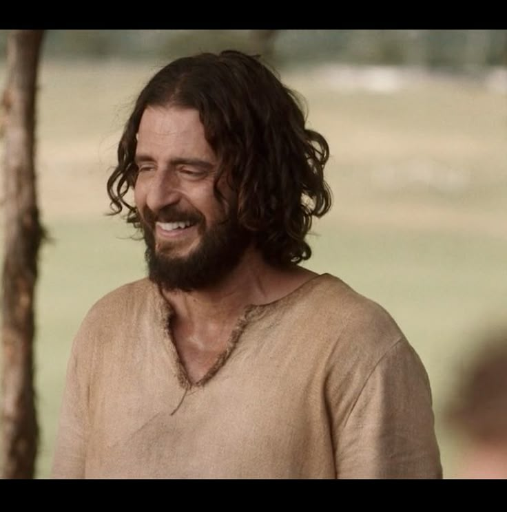 a man with long hair and a beard smiles at the camera while standing next to a tree