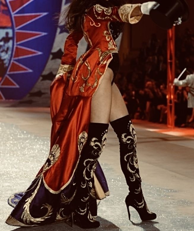 a woman is walking down the runway wearing high heeled boots and a red dress