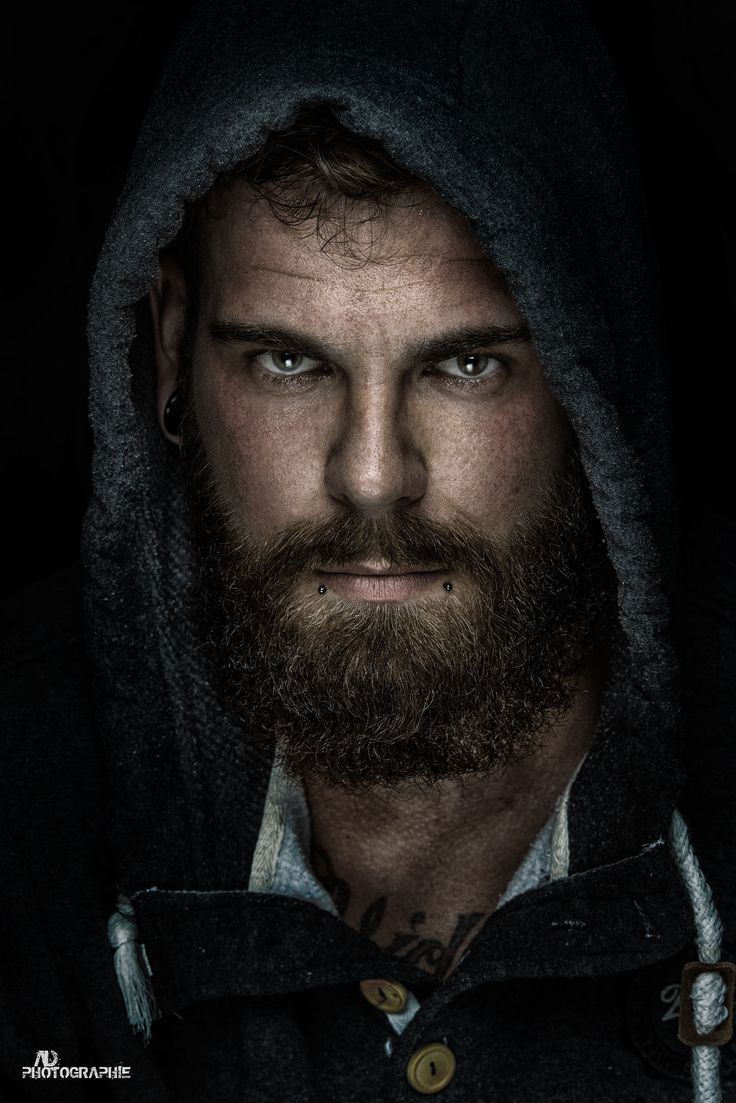 a man with a beard wearing a hoodie and looking at the camera while standing in front of a dark background