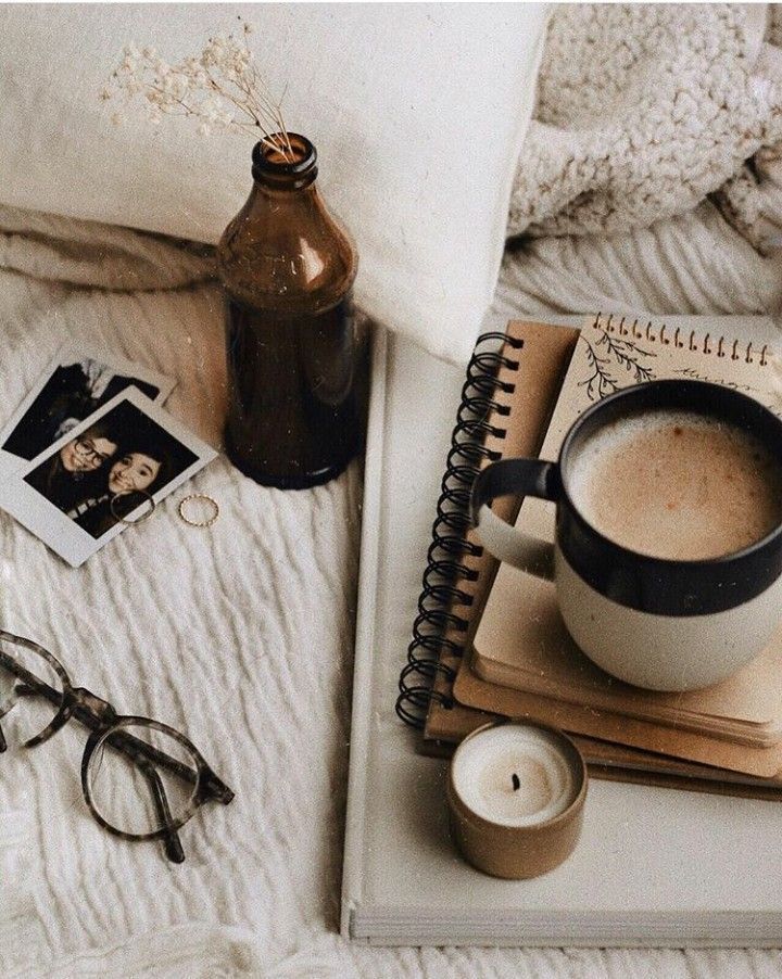 a cup of coffee on a bed next to a book and eyeglasses with an open notebook