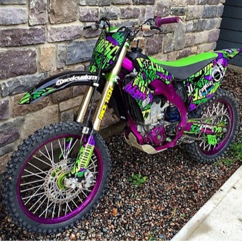 a purple and green dirt bike parked next to a brick wall with graffiti on it