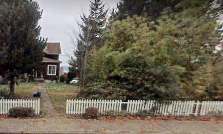 a white picket fence in front of a house with trees and bushes around it on a cloudy day