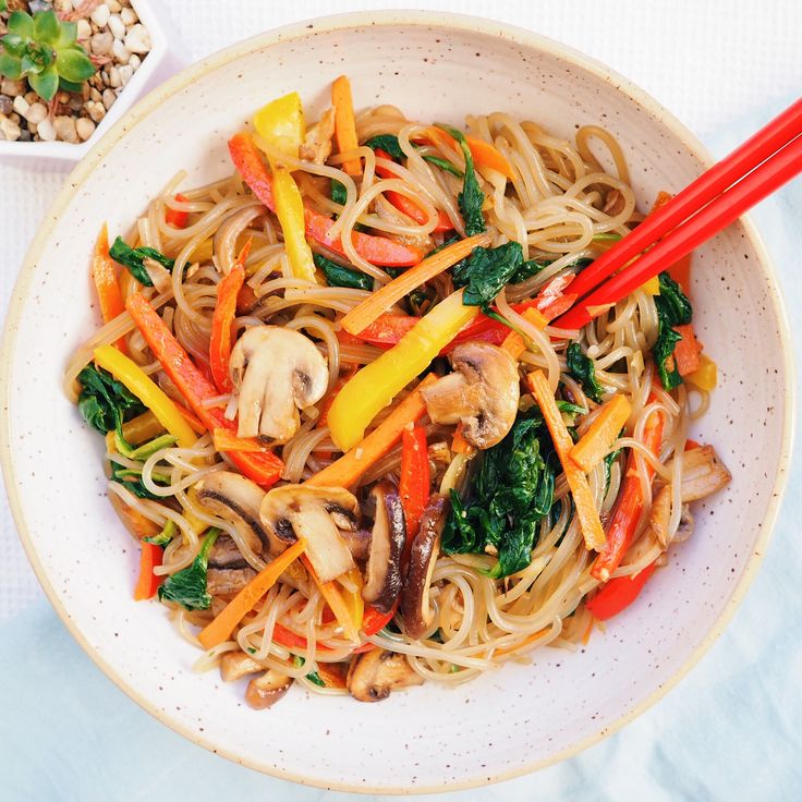 a white bowl filled with noodles and vegetables next to chopsticks on a table