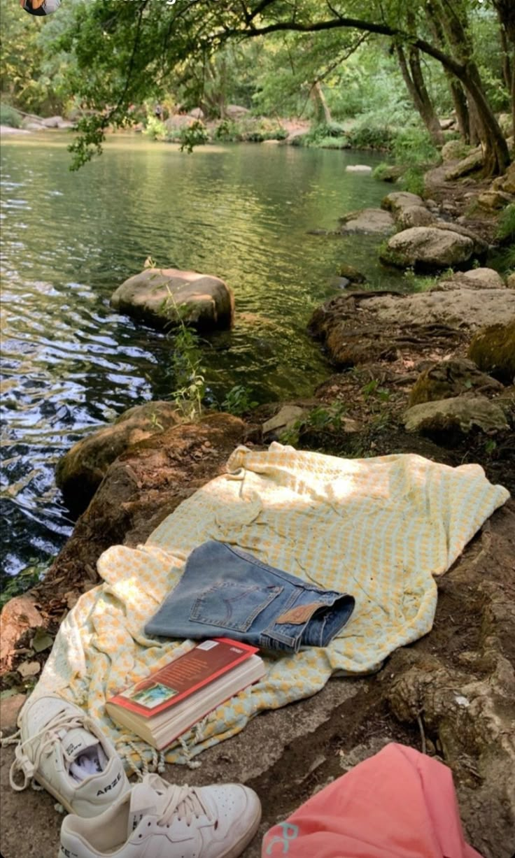 an open book and some books on a blanket by the water with trees in the background